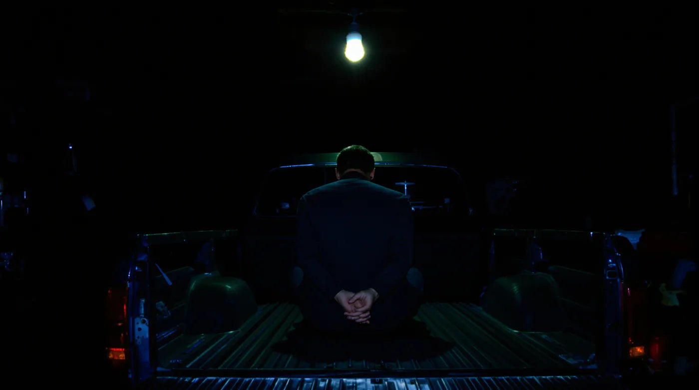A man sitting alone in the back of a pickup truck in a dark garage, head down.