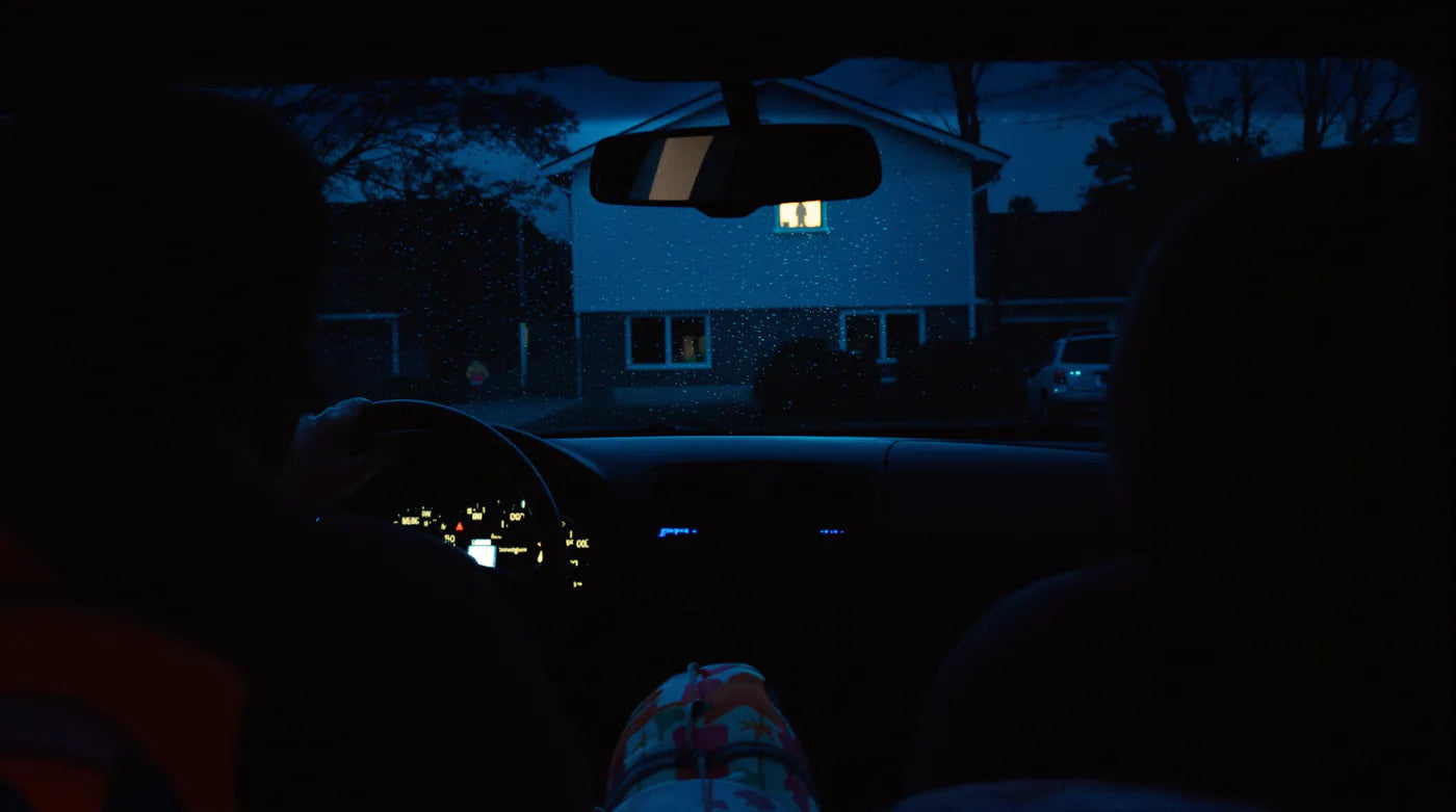 Night, inside a parked car: a child watches from a lit upstairs window. A small backpack was left on the seat.