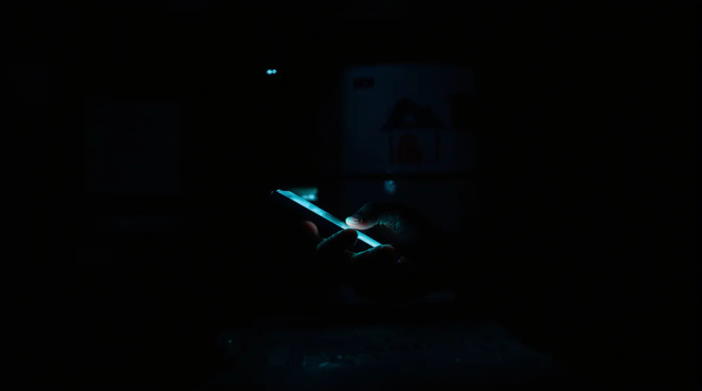 A dark kitchen at night, a man's hand lit by a phone screen, a child's drawing of a house on the fridge.