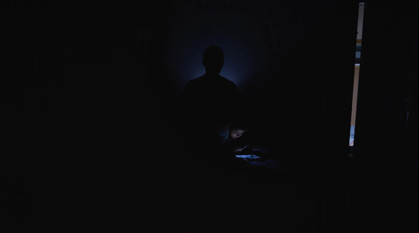 A child lit by a tablet in a dark bedroom, a father's silhouette behind.